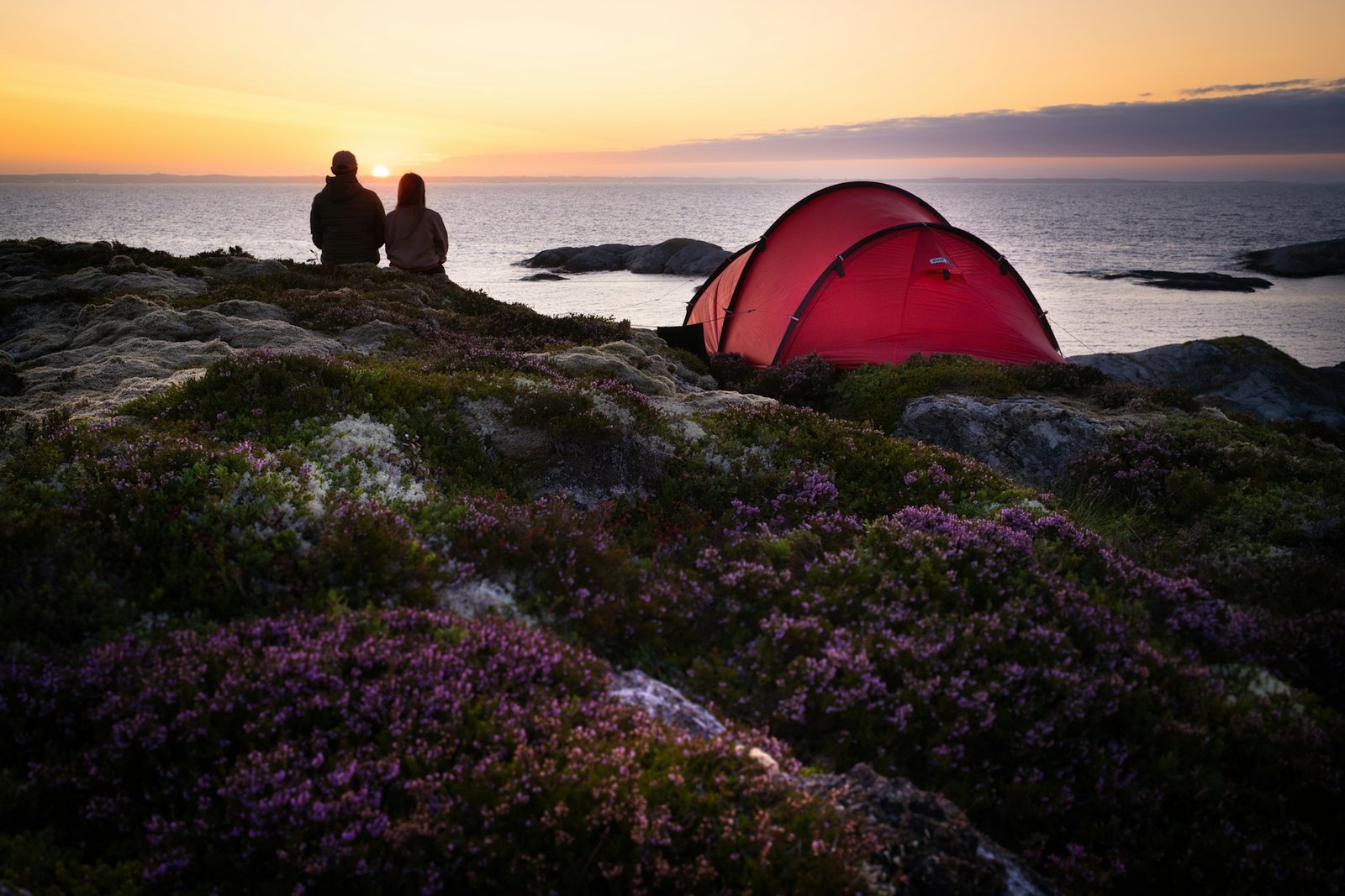 A couple sitting close together on coastal rocks beside a red tent, watching the sunset over the sea