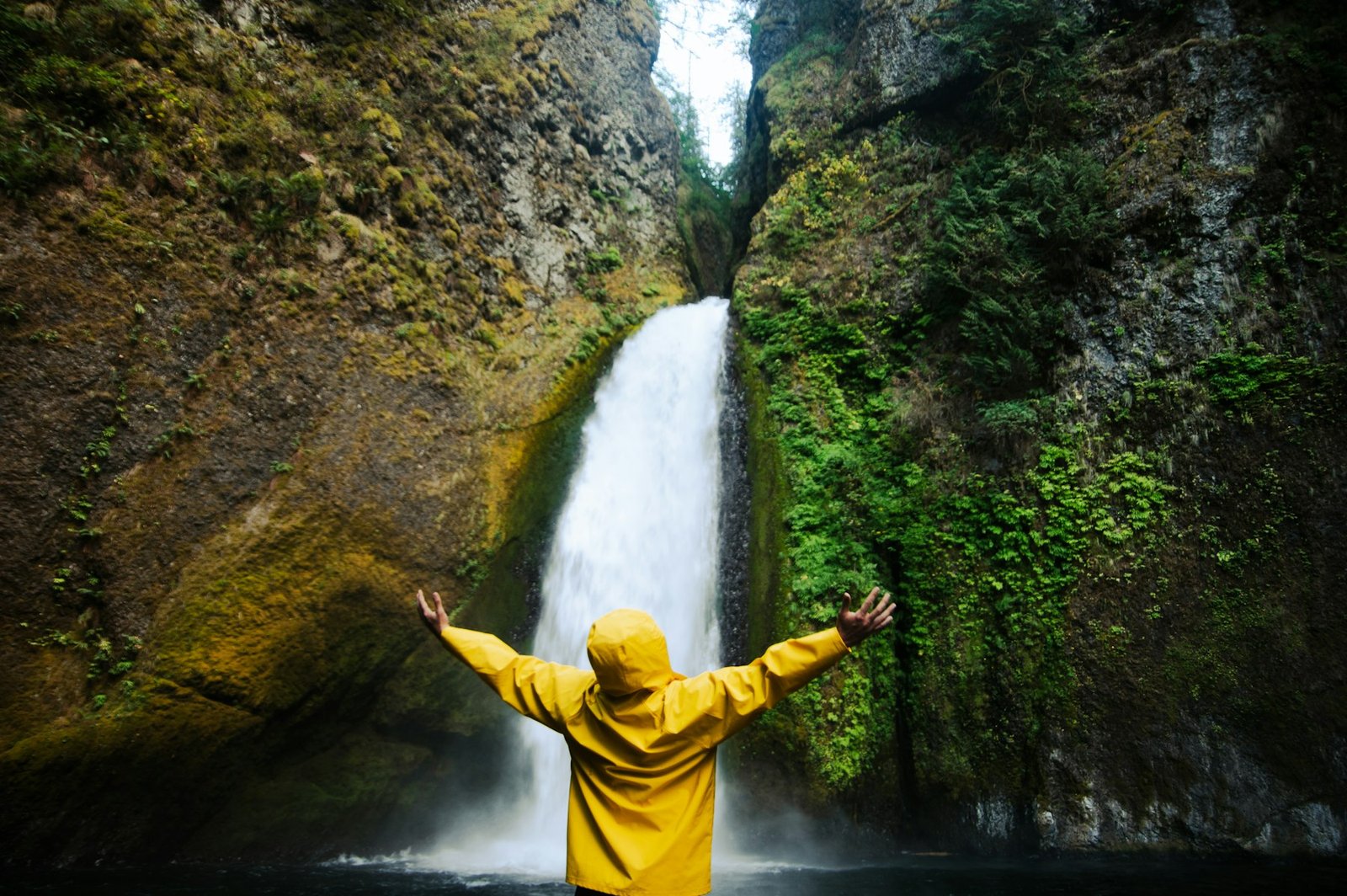 Person in a yellow raincoat looking up at a tall waterfall